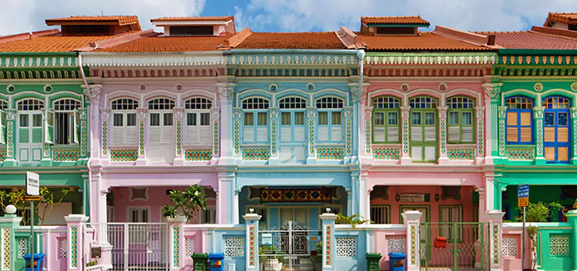 Row of colorful Peranakan shophouses with ornate facades and pastel tones, showcasing traditional architecture under a bright blue sky.
