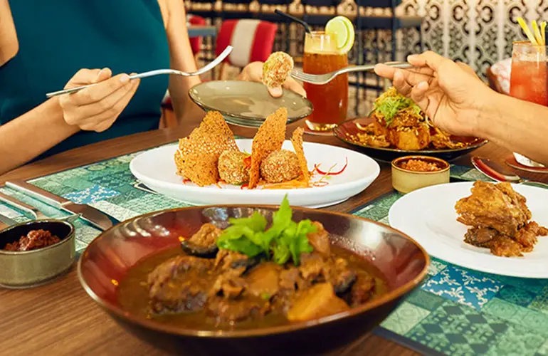 Close-up of a vibrant Peranakan meal with richly spiced dishes, colorful drinks, and diners enjoying food at a beautifully set restaurant table.