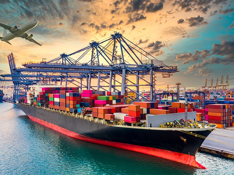 Cargo ship loaded with colorful containers docked at a busy port with cranes, under a dramatic sunset sky, and an airplane flying overhead.