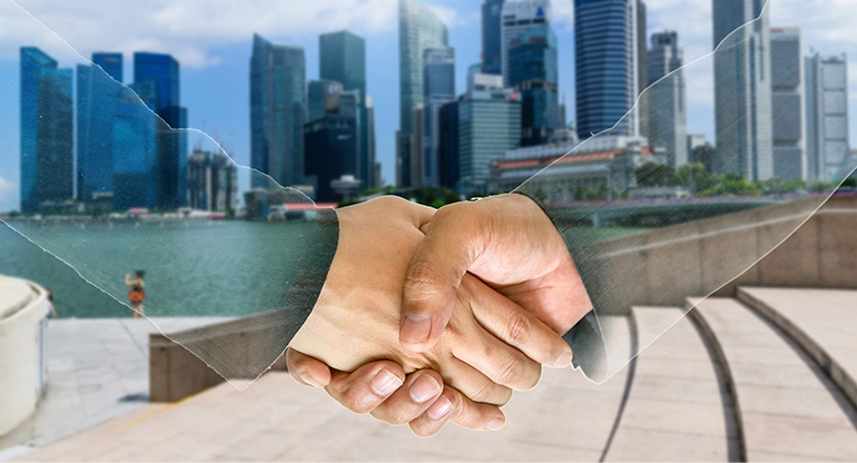 Two hands shaking in the foreground with a transparent overlay effect, set against the backdrop of Singapore’s Marina Bay skyline, symbolizing partnership and business collaboration.