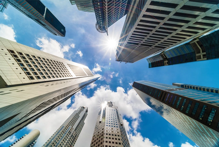 Upward view of modern skyscrapers against a bright blue sky with scattered clouds, capturing sunlight reflecting off the glass buildings.