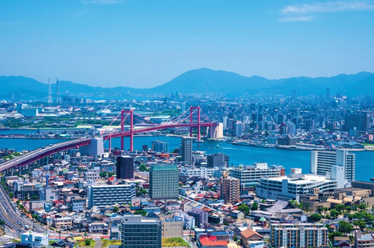 Cityscape featuring a red suspension bridge spanning over a bay, surrounded by urban buildings, with distant mountains under a clear blue sky.