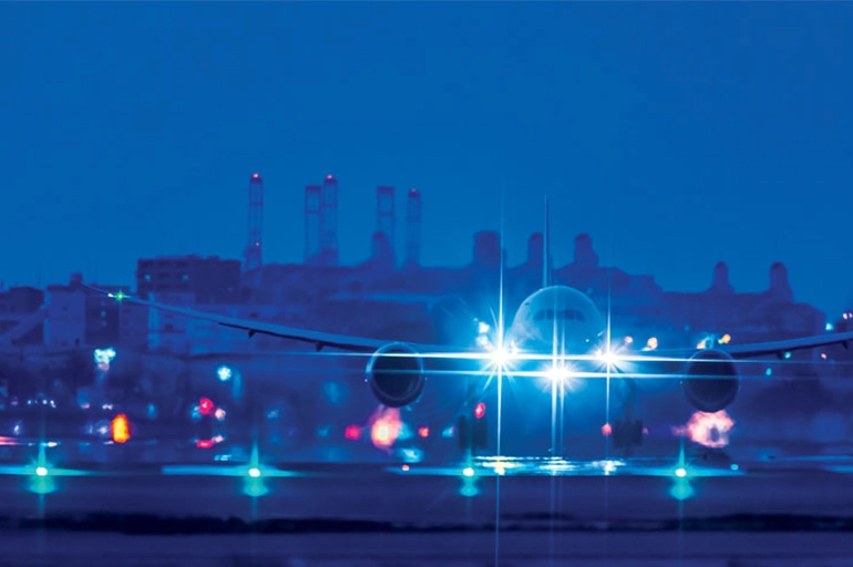 Airplane with bright landing lights preparing for takeoff on a runway at night, with a city skyline and industrial structures visible in the background.