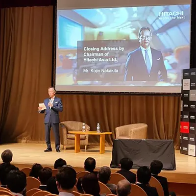 Chairman of Hitachi Asia Ltd., Mr. Kojin Nakakita, delivering the closing address on stage during a corporate event, with an audience seated in front and a presentation screen behind him.