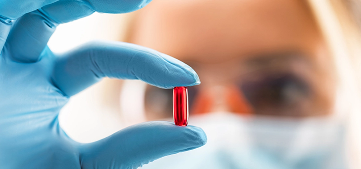 Close-up of a gloved hand holding a red capsule pill, with a masked scientist or lab technician examining it in the background.