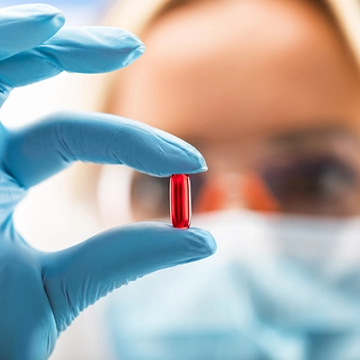 Close-up of a gloved hand holding a red capsule pill, with a masked scientist or lab technician examining it in the background.