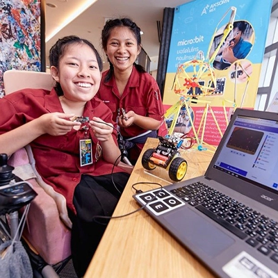 Two smiling students in red uniforms showcasing a robotics project with a laptop, a wheeled robot, and a micro:bit display at a tech exhibition.