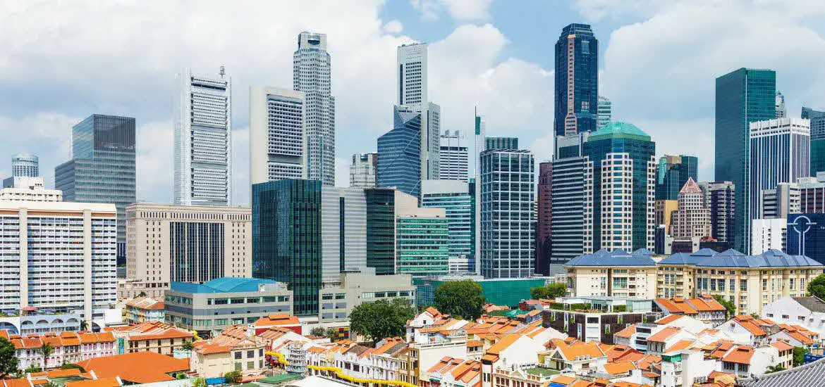 A vibrant view of Singapore’s central business district showcasing a dense cluster of modern skyscrapers contrasted with the traditional shophouses featuring red-tiled roofs in the foreground under a partly cloudy sky.