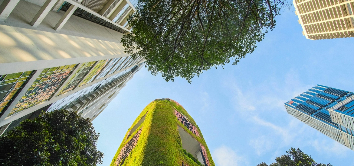 Upward view of modern high-rise buildings with greenery-covered facades and surrounding trees against a clear blue sky.