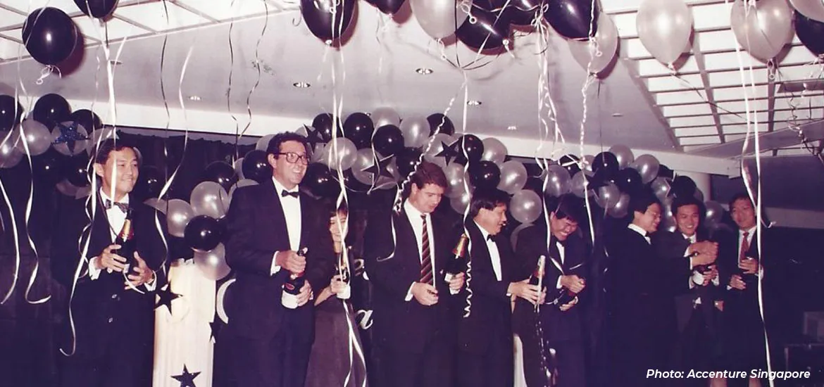 Group of men in formal suits holding champagne bottles at a celebration, with black and silver balloons and streamers decorating the room.