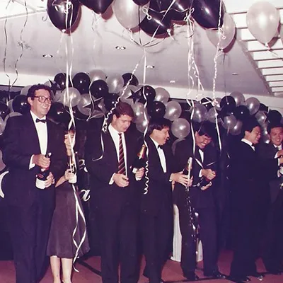 Group of men in formal suits holding champagne bottles at a celebration, with black and silver balloons and streamers decorating the room.