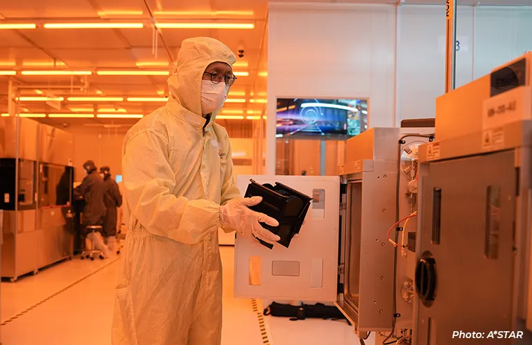 A technician in a cleanroom suit operating semiconductor manufacturing equipment, carefully handling a component inside a controlled lab environment.