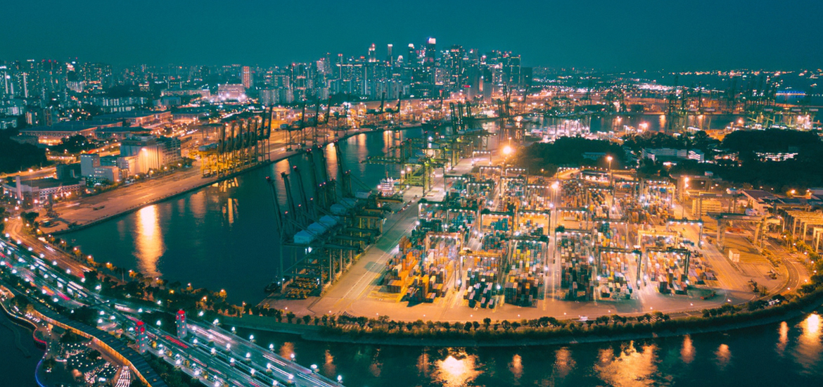 Aerial nighttime view of Singapore’s container port and waterfront logistics hub, with brightly lit shipping terminals, cargo cranes, container yards, and the city skyline in the background.