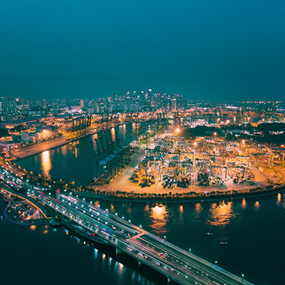 Aerial nighttime view of Singapore’s container port and waterfront logistics hub, with brightly lit shipping terminals, cargo cranes, container yards, and the city skyline in the background.