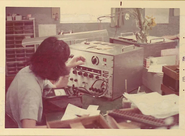 Vintage photograph of a technician operating and adjusting electronic testing equipment at a workbench, surrounded by documents, instruments, and laboratory tools in an early electronics development or manufacturing environment.