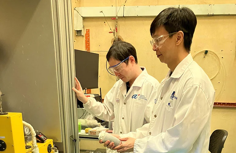 Two researchers in laboratory coats and safety goggles examining a test sample beside scientific equipment, conducting materials research and development in a laboratory environment.