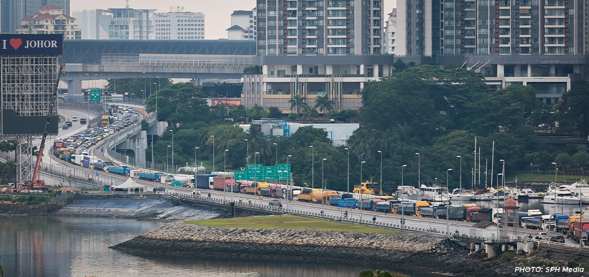 Heavy traffic congestion along the Johor–Singapore Causeway with trucks and vehicles entering Johor Bahru, against a backdrop of high-rise buildings and waterfront areas.