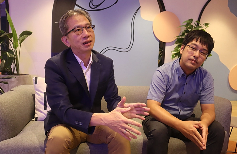 Two men seated on a sofa in a casual office setting, engaged in discussion, with one speaking and gesturing while the other listens.