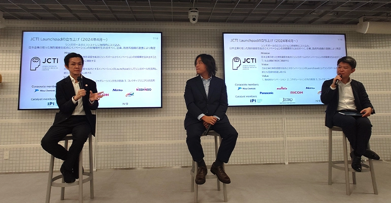 Panel discussion with three speakers seated on stools, speaking into microphones in front of screens displaying information about the JCTI launch and partner organizations.