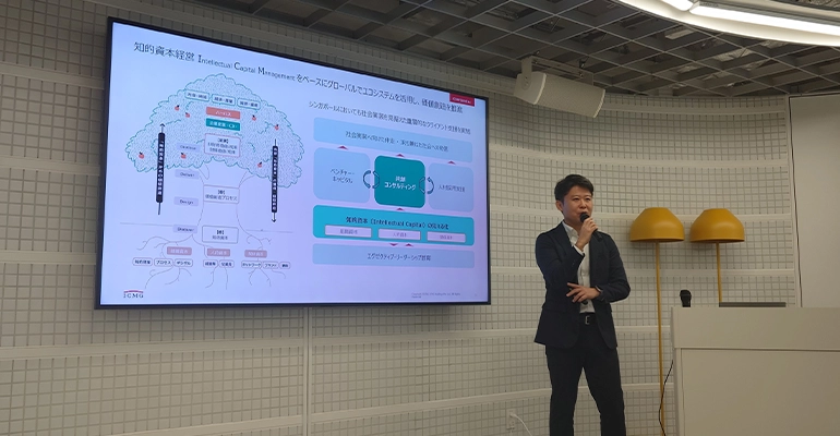 Panel discussion with three speakers seated on stools, speaking into microphones in front of screens displaying information about the JCTI launch and partner organizations.