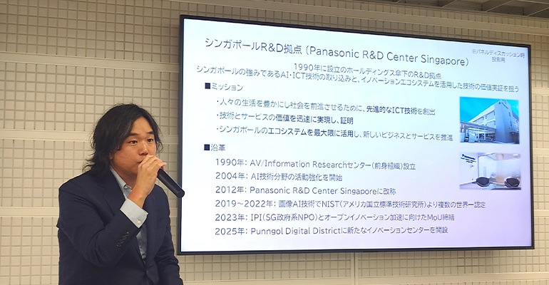 Panel discussion with three speakers seated on stools, speaking into microphones in front of screens displaying information about the JCTI launch and partner organizations.