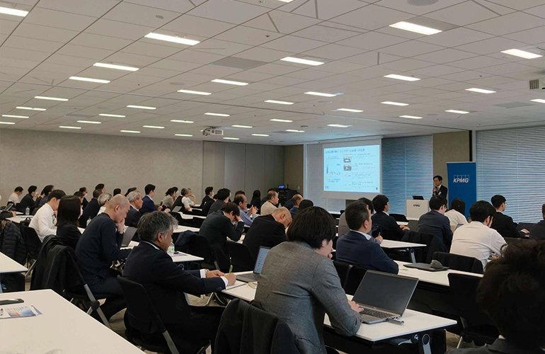 Audience of professionals seated in a conference room attending a presentation, with a speaker presenting slides at the front.