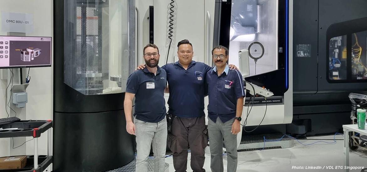 Three engineers standing together and smiling inside a modern manufacturing facility with advanced machinery and equipment in the background.
