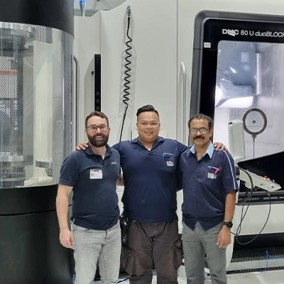 Three engineers standing together and smiling inside a modern manufacturing facility with advanced machinery and equipment in the background.