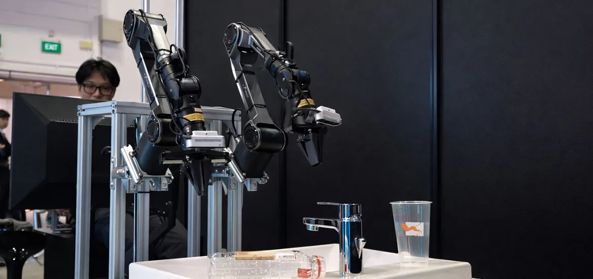 Two robotic arms mounted on a frame positioned over a sink with a faucet, bottle, and plastic cup, while a person observes from behind.