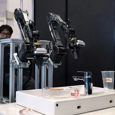 Two robotic arms mounted on a frame positioned over a sink with a faucet, bottle, and plastic cup, while a person observes from behind.