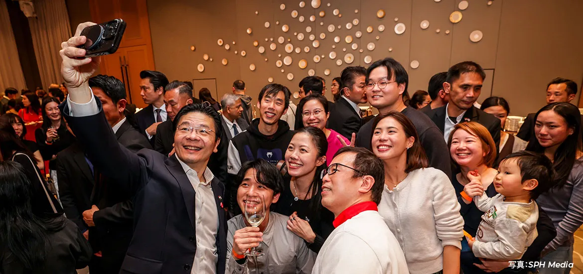 A group of people at a social event gathering closely together to take a selfie, smiling and posing, with one person holding up a camera while others—including a child—join in.