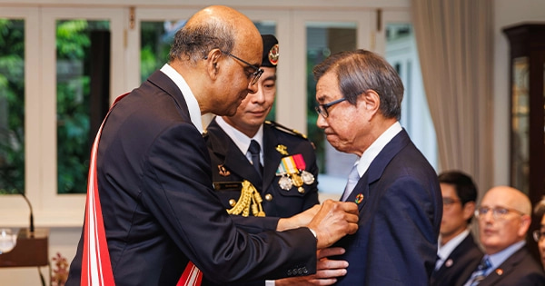 Singapore President Tharman Shanmugaratnam presents a national medal to an elderly man in a formal ceremony, with uniformed personnel in attendance.