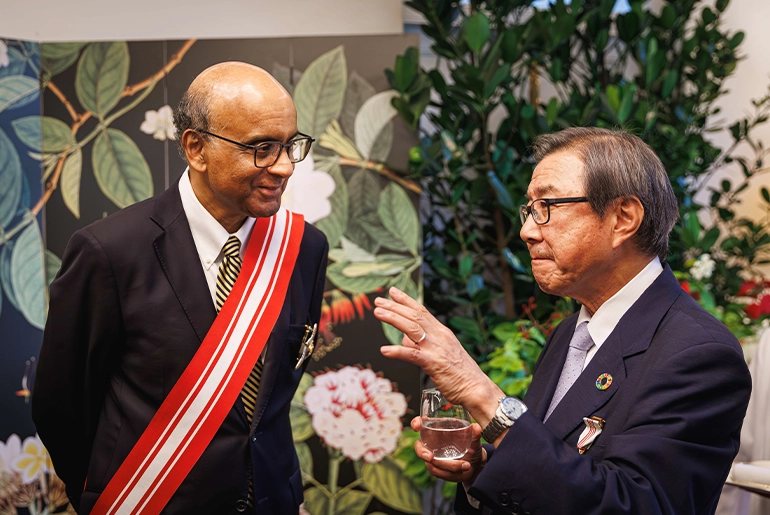 Singapore President Tharman Shanmugaratnam converses with an elderly medal recipient in a formal setting adorned with floral decor and greenery. Both men are dressed in suits, with visible medals of distinction.