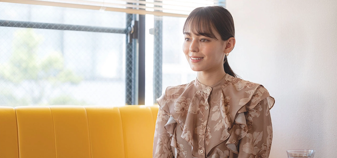 Smiling woman in a floral blouse sitting on a yellow bench in a well-lit room with large windows and soft natural light.