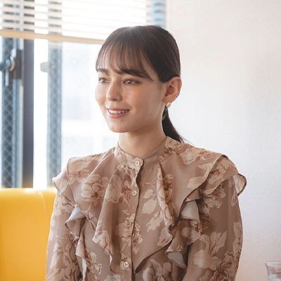 Smiling woman in a floral blouse sitting on a yellow bench in a well-lit room with large windows and soft natural light.