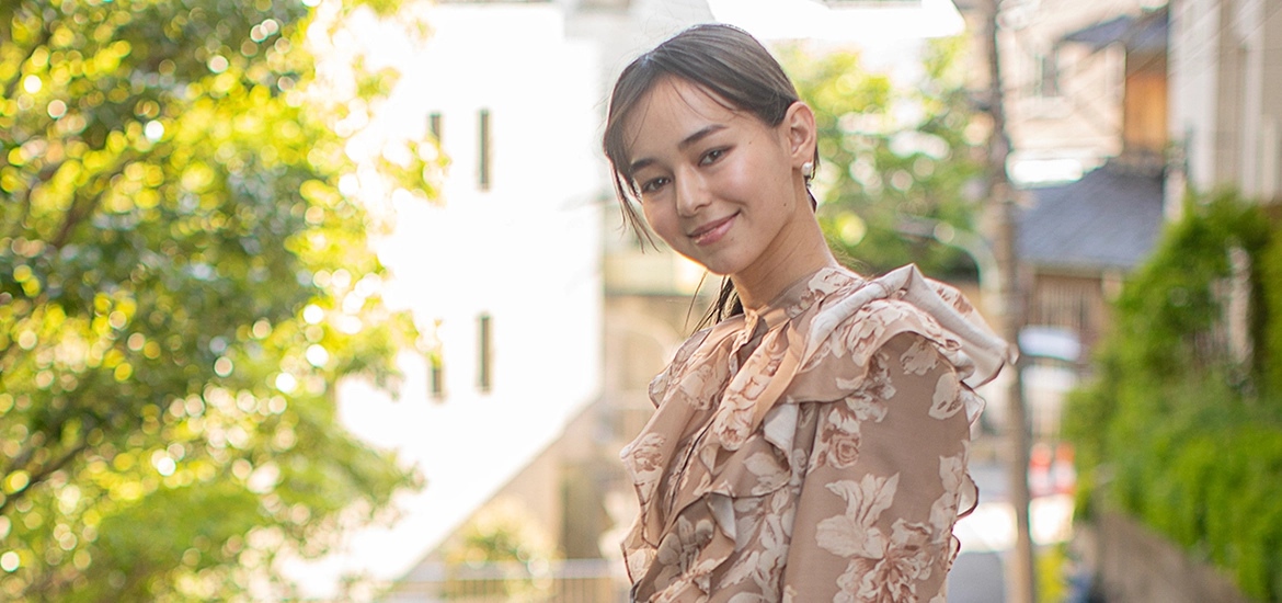 Smiling woman in a floral blouse standing outdoors on a sunlit street, with greenery and soft-focus buildings in the background.