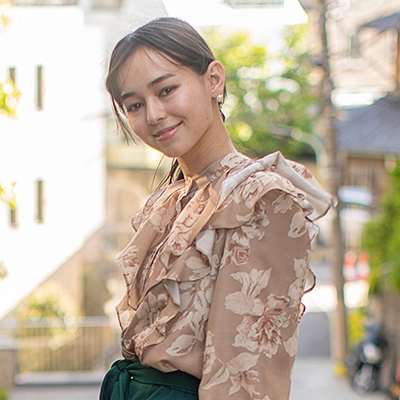Smiling woman in a floral blouse standing outdoors on a sunlit street, with greenery and soft-focus buildings in the background.