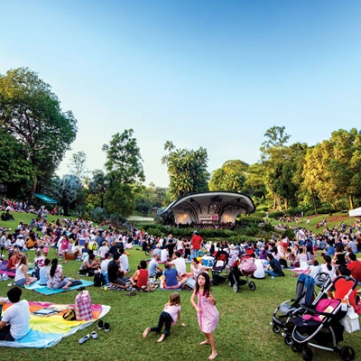Large crowd enjoying an outdoor concert at a lush green park, with families and friends sitting on picnic mats facing a modern stage surrounded by trees.