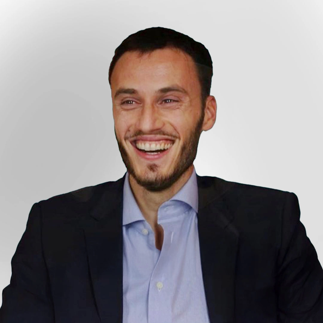 A smiling man in business attire, wearing a blazer and open-collar shirt, against a plain background.