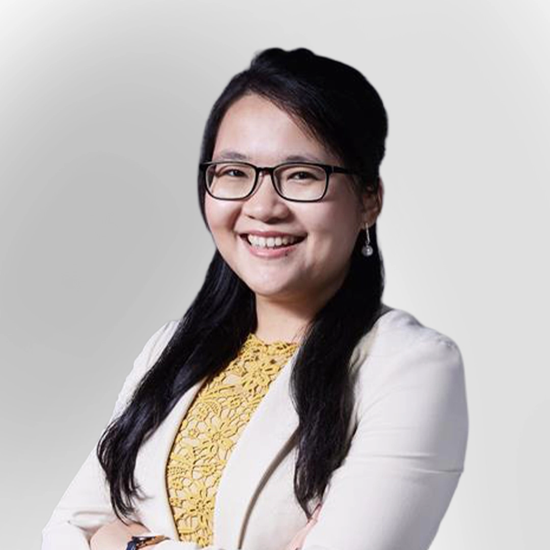 Portrait of a woman wearing glasses, a light blazer, and a yellow top, smiling with arms crossed against a plain background.